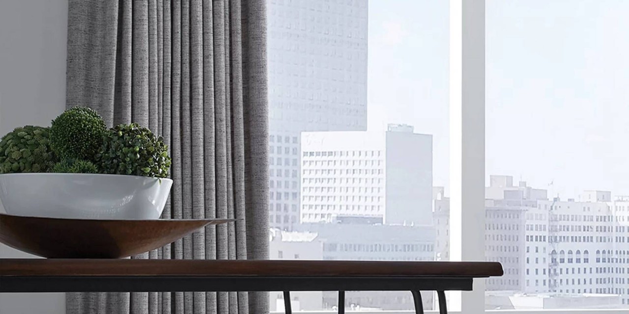 Small table featuring a bowl of green plants set in front a large windows with Carole Fabrics custom drapes looking out into the city. 